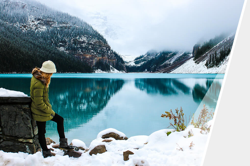 Eine Person in Winterkleidung steht nachdenklich am Rand eines kristallklaren Bergsees, umgeben von schneebedeckten Bergen, wobei die ruhige Szene die majestätische Schönheit der Natur widerspiegelt.